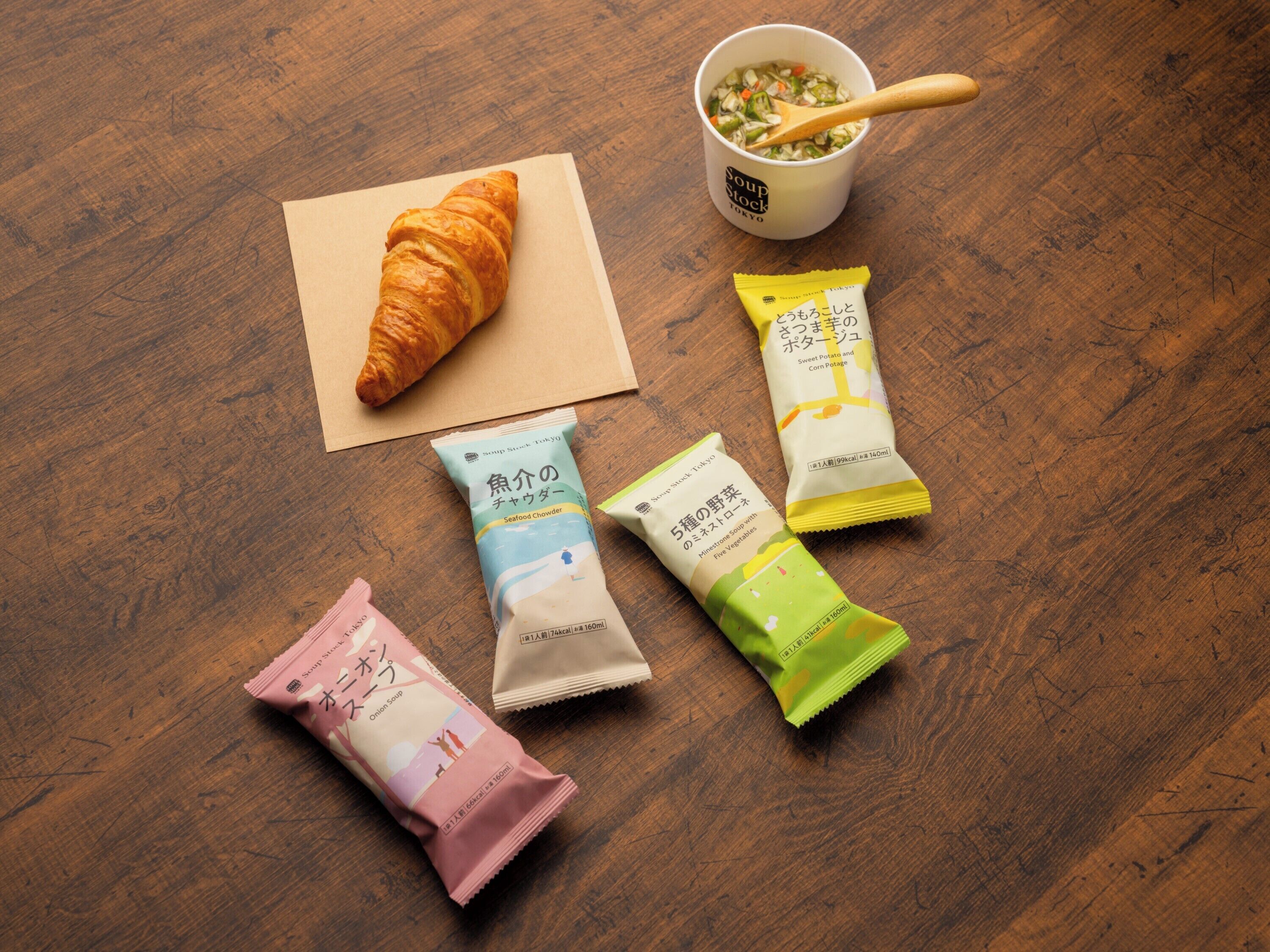 A croissant, a cup of soup, and four packets of soup on a wooden table.