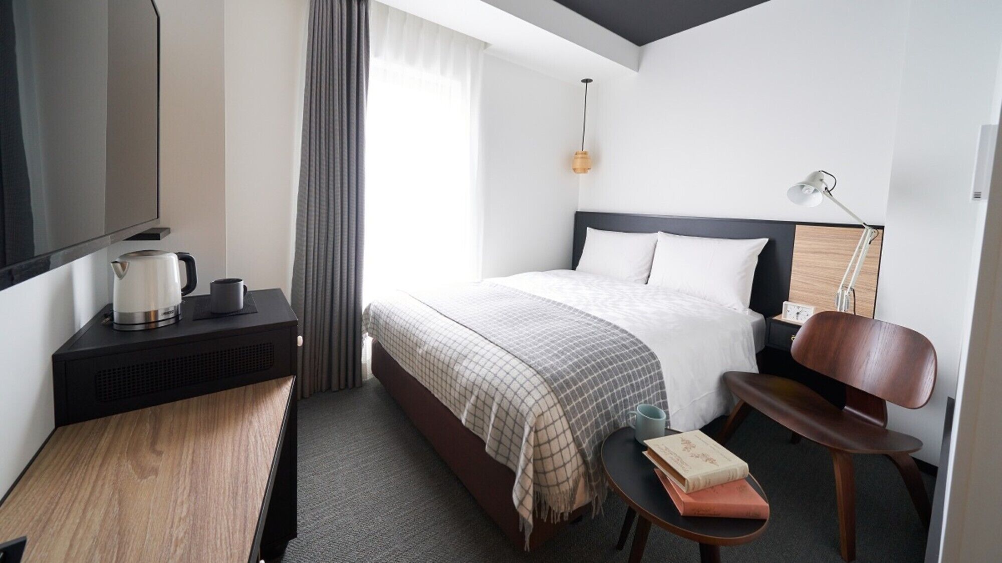A hotel room with a bed, bedside table, chair, lamp, and a small table with books.