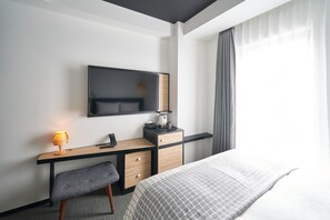 Quarto de hotel moderno com cama, escrivaninha com cadeira, luminária de mesa, TV de tela plana e mesa de cabeceira com abajur.