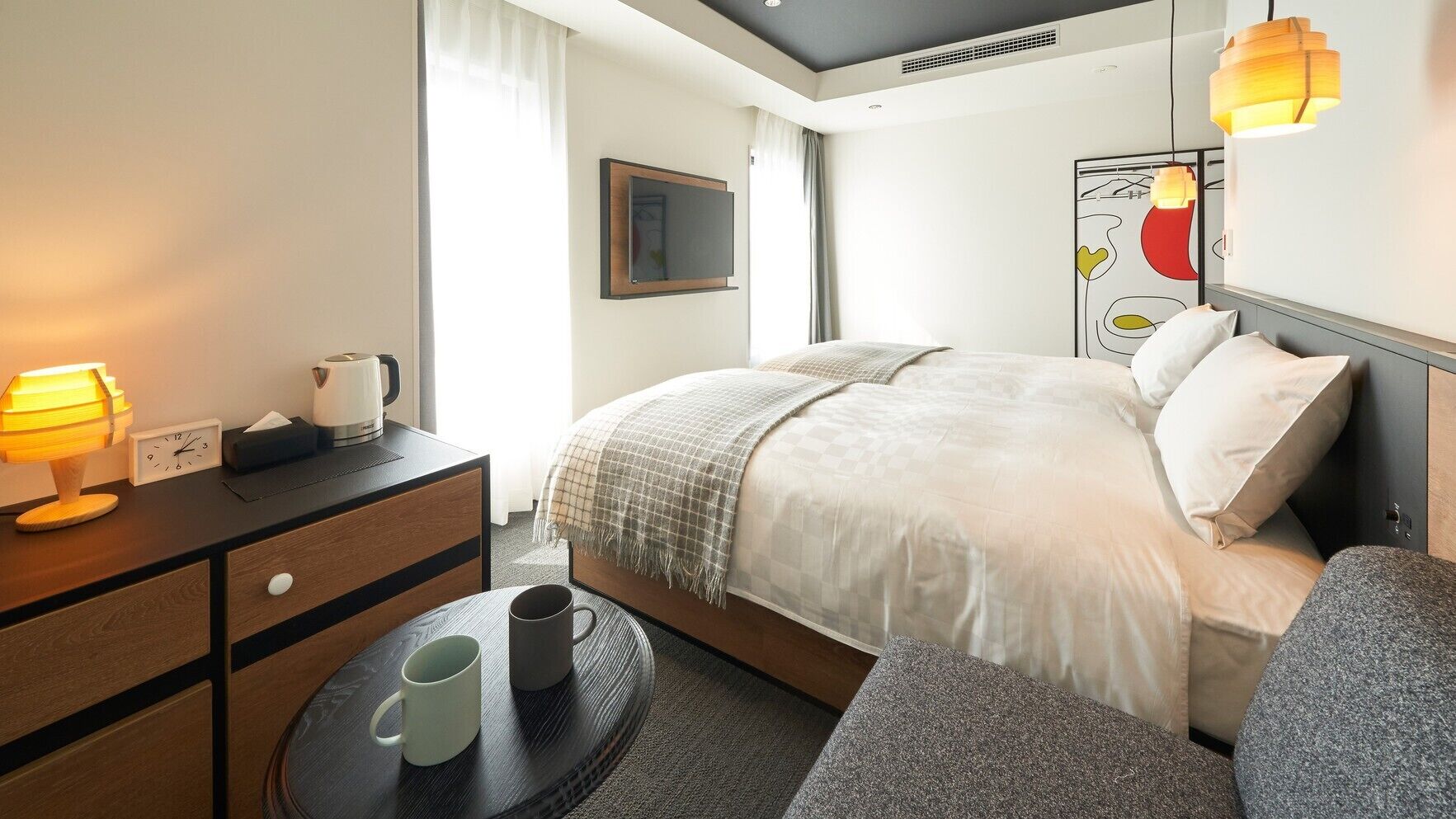A hotel room with a bed, bedside table, lamp, clock, and a small table with a mug.