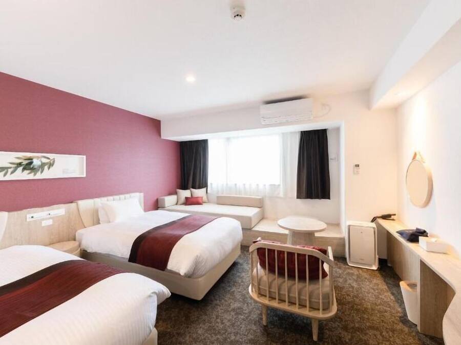 A hotel room with two beds, a small round table, a chair, a desk with a flat-screen TV, and a wall-mounted air conditioner.