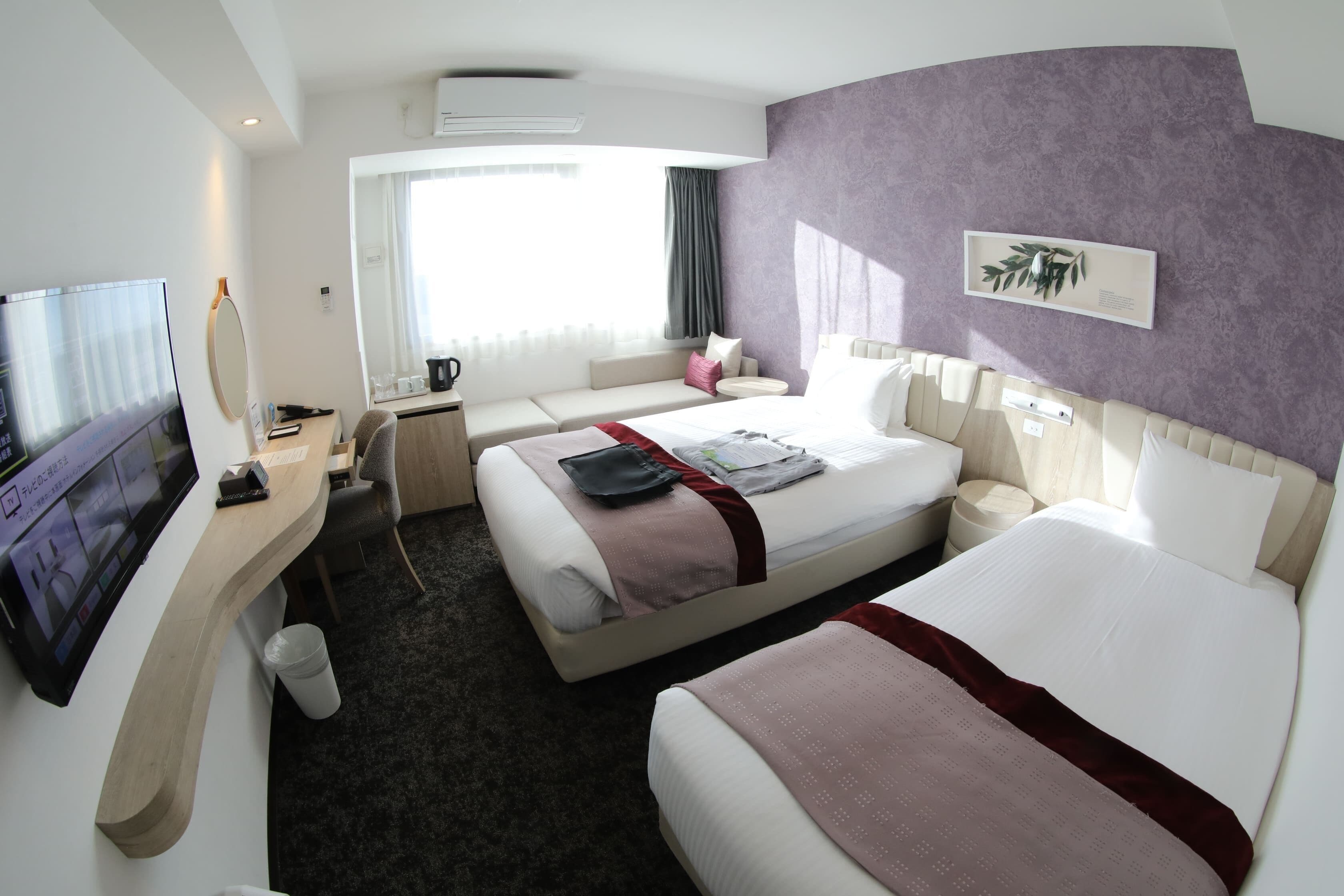 Ein Hotelzimmer mit zwei Betten, einem Schreibtisch mit Fernseher, einem Fenster mit Vorhängen und einer Wandpflanze.