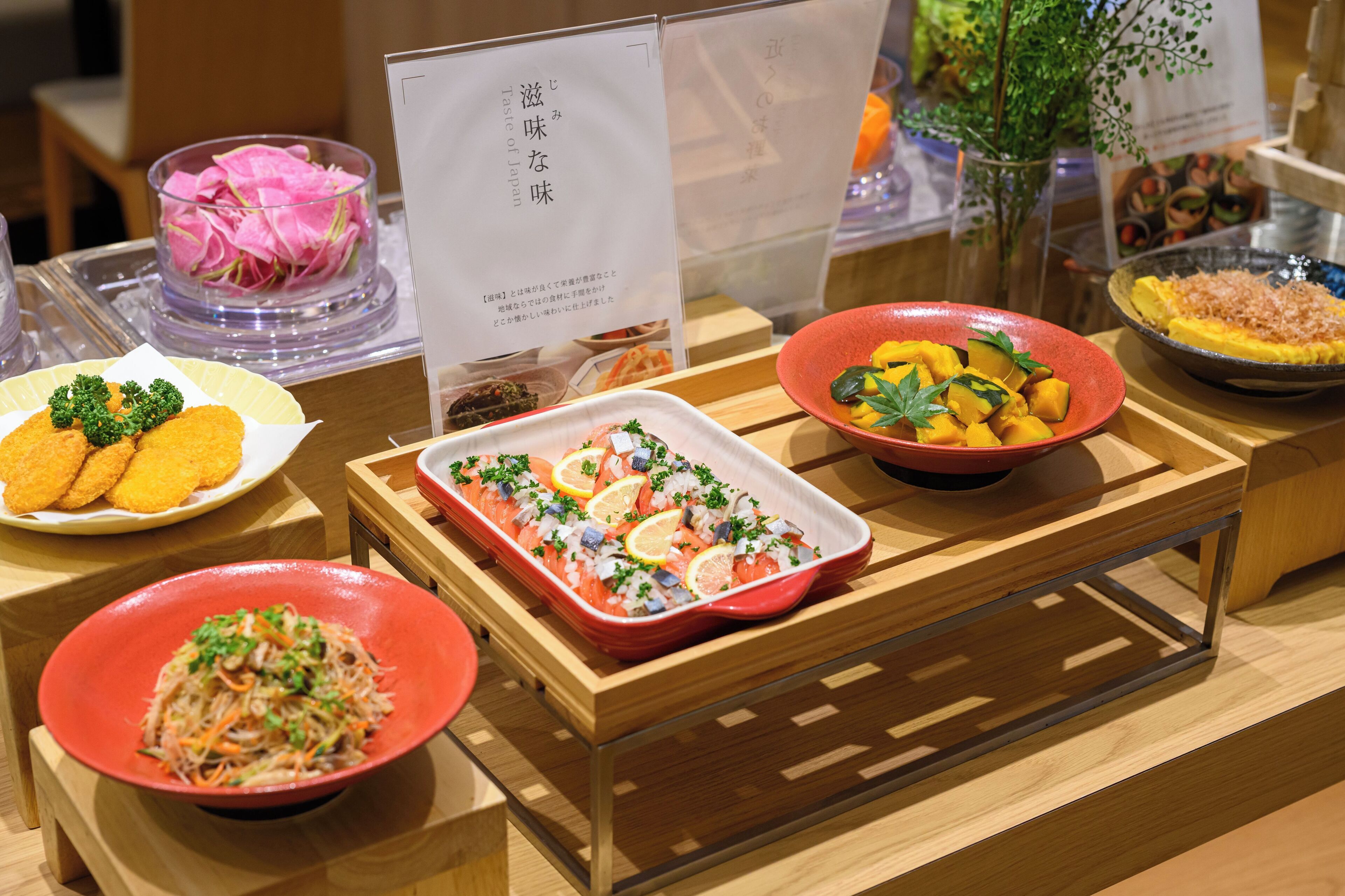 A variety of dishes displayed on a wooden table, including a dish with orange slices and herbs, a dish with yellow vegetables, and a dish with a red bowl.