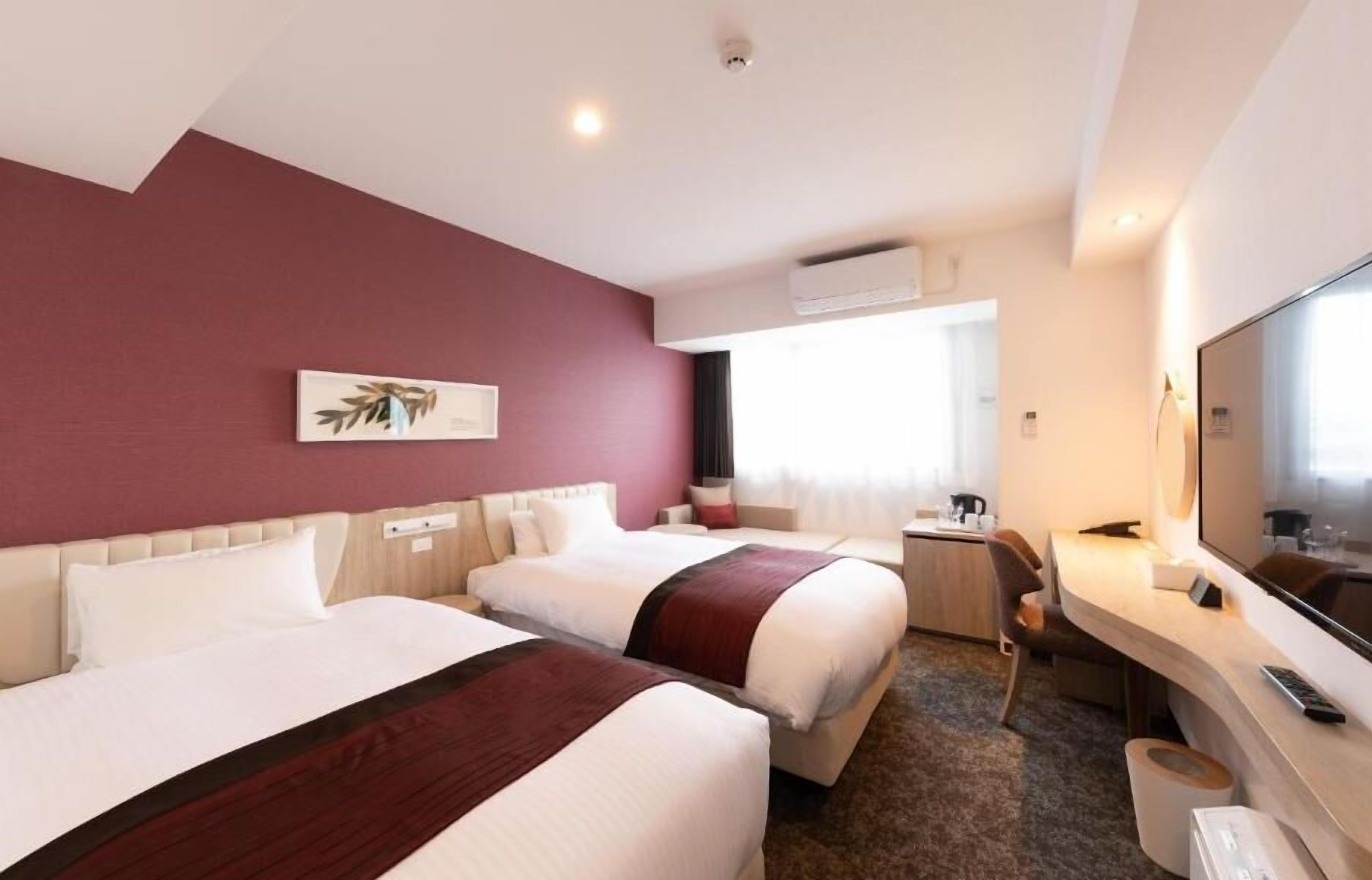 Ein Hotelzimmer mit zwei Betten, einem Schreibtisch, einem Fernseher und einem Fenster.