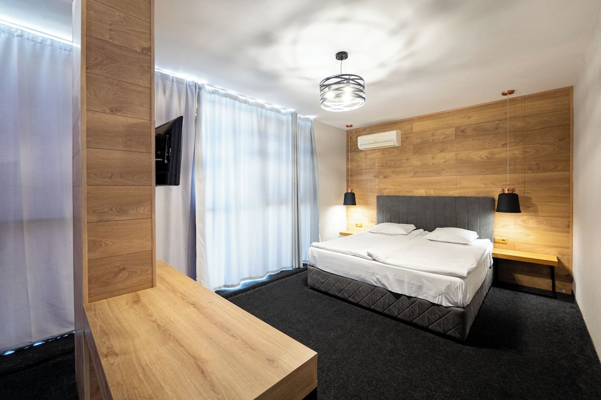 A modern hotel room with a wooden headboard, a bed with white linens, a bedside table, and a wall-mounted TV.