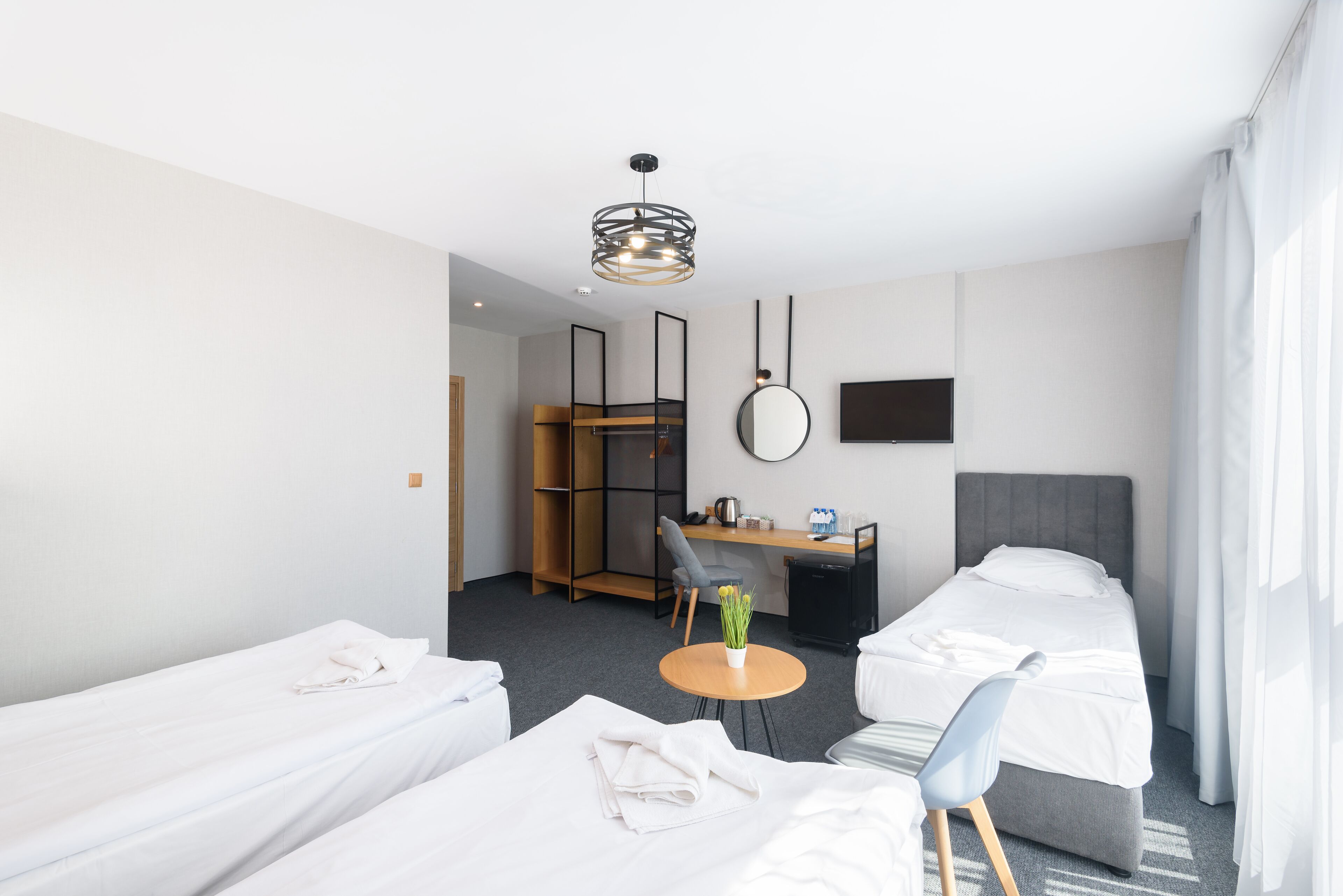 A modern hotel room with two single beds, a desk with a chair, a small round table with a vase, a TV mounted on the wall, and a ceiling light fixture.