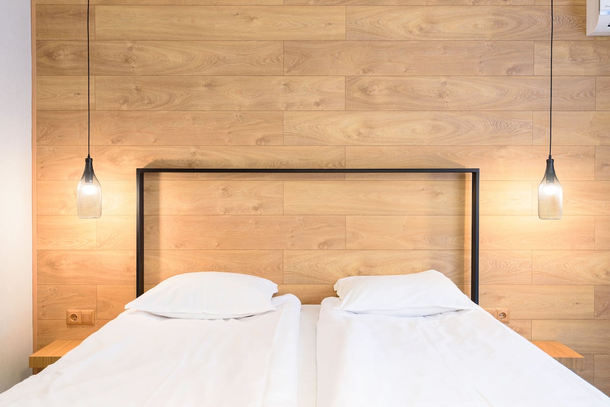 A double bed with white linens, a wooden headboard, and two pendant lights.