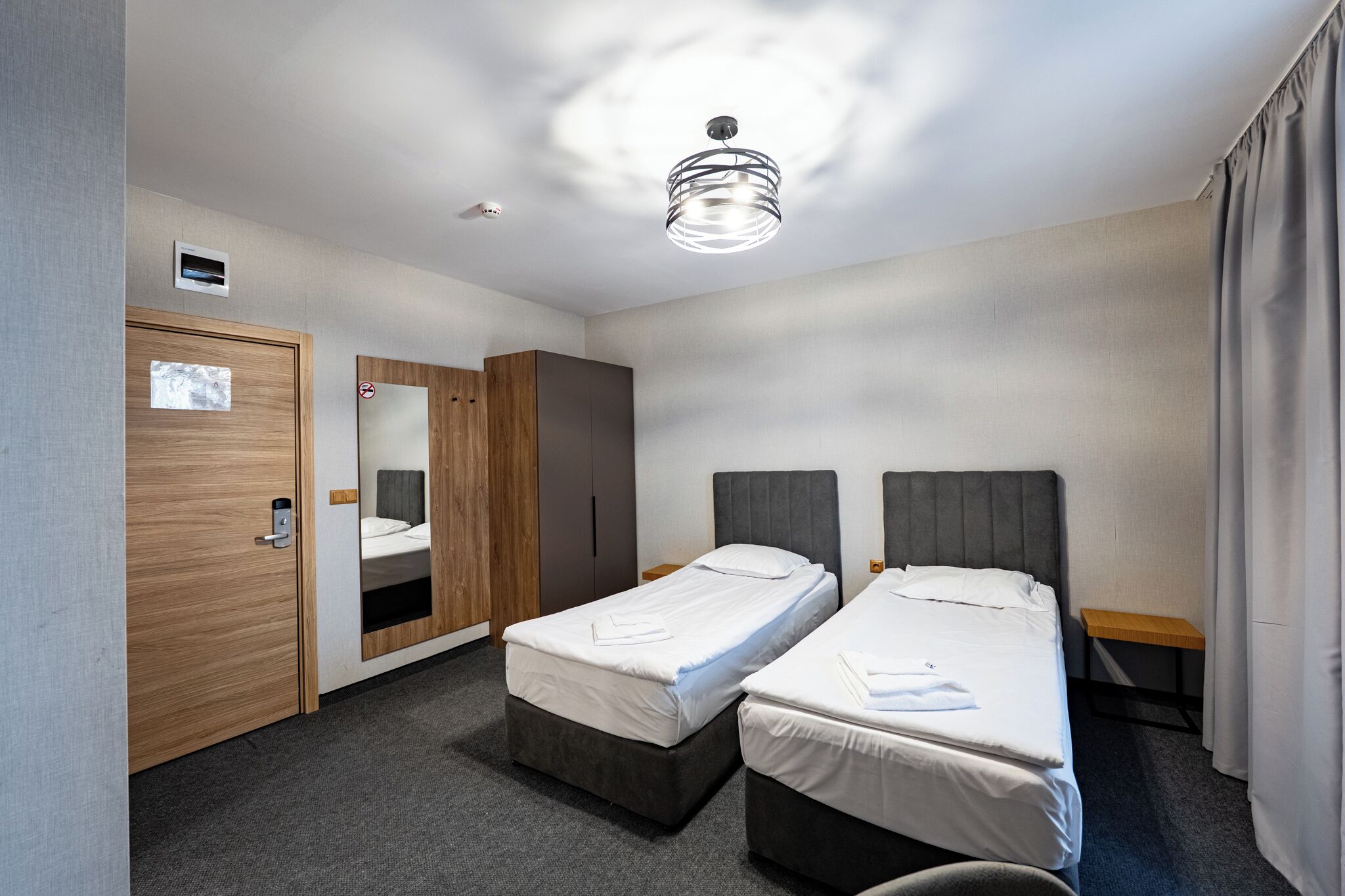A hotel room with two single beds, a wooden wardrobe, a mirror, a ceiling light fixture, and a small wooden nightstand.