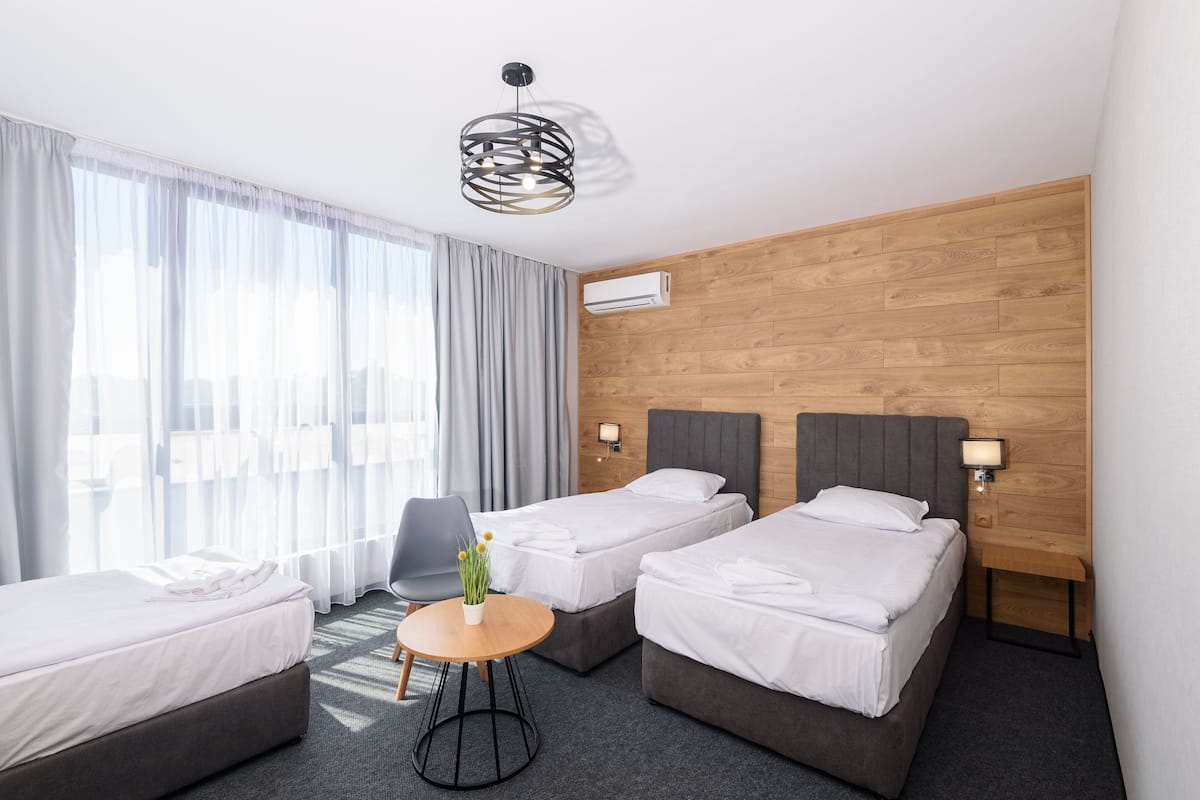A hotel room with two single beds, a small round table with a vase of flowers, a grey armchair, and a modern pendant light.