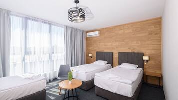 A hotel room with two single beds, a small round table with a vase of flowers, a grey armchair, and a modern pendant light.