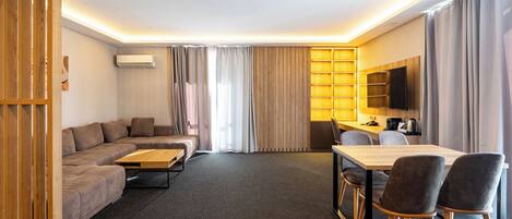 A modern hotel room with a sofa, a dining table with chairs, a TV, and a desk.