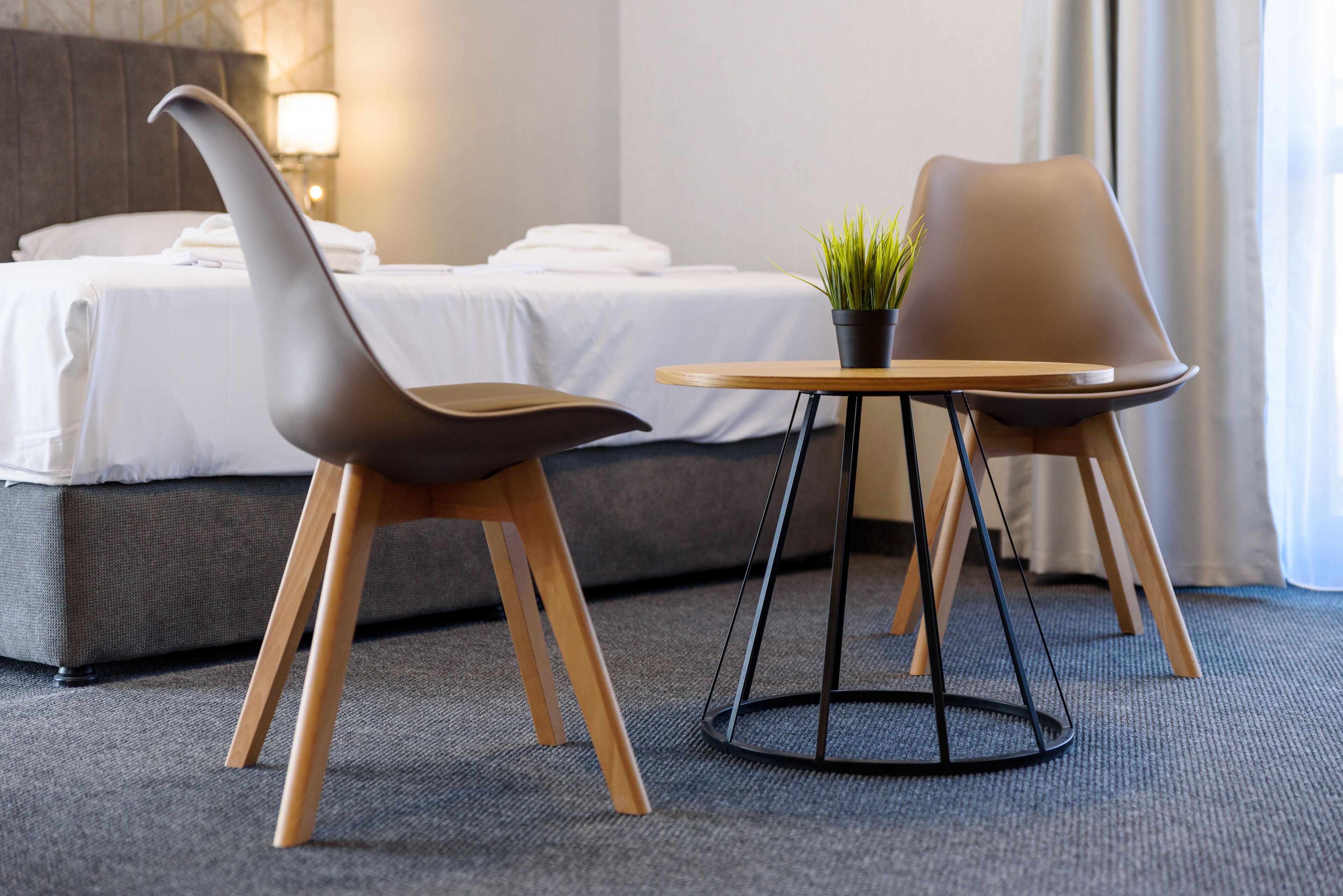 Habitación de hotel con cama, dos sillas modernas, una mesita redonda con planta en maceta y una lámpara.