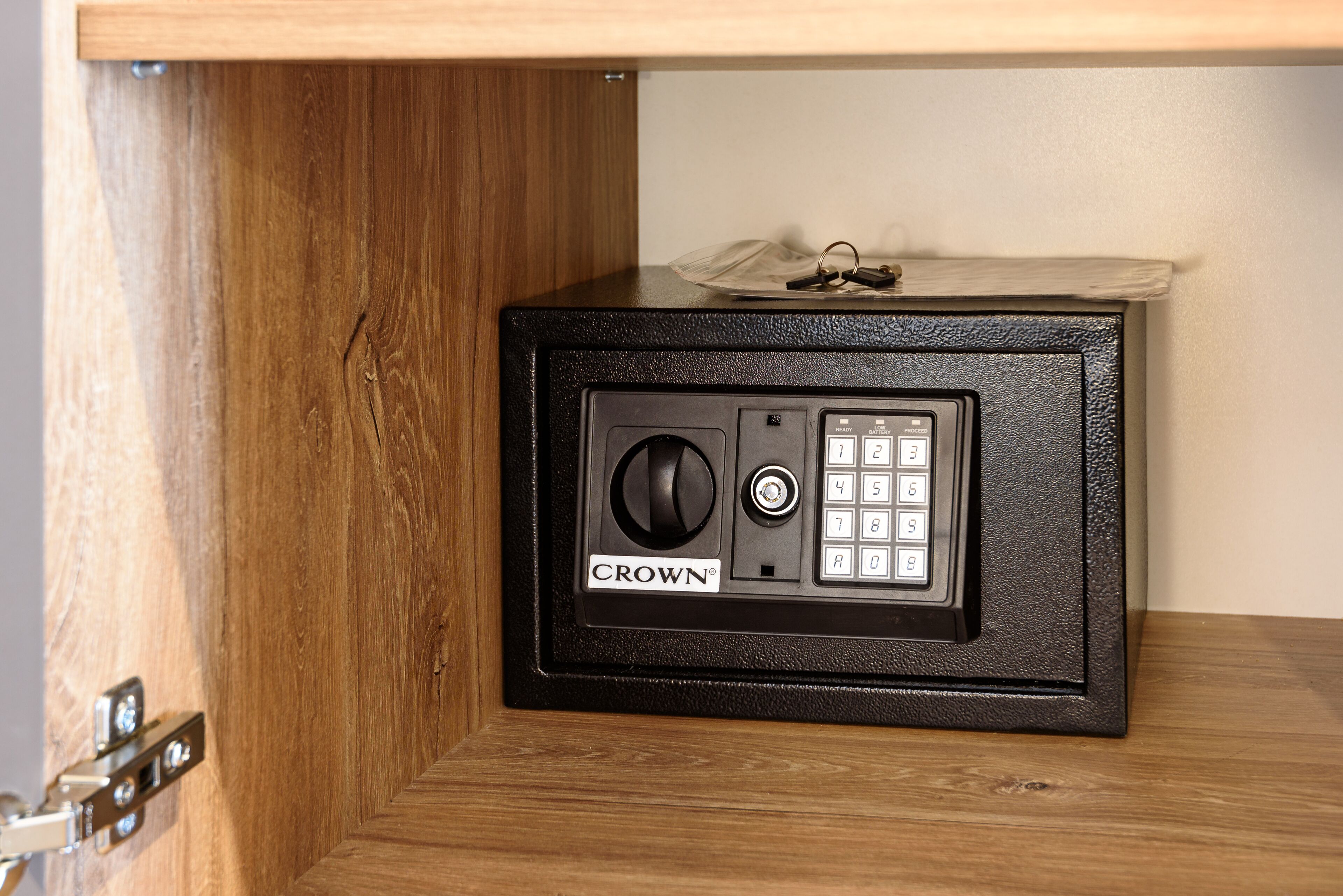 Una caja fuerte con teclado y cerradura de combinación, identificada con la etiqueta “CROWN”, ubicada debajo de un estante de madera.