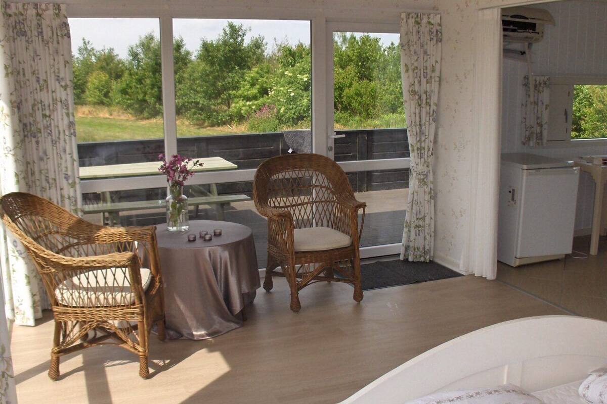 Ein Schlafzimmer mit einem Bett, zwei Korbstühlen, einem kleinen runden Tisch mit Blumenvase und Blick auf Grün durch große Fenster.
