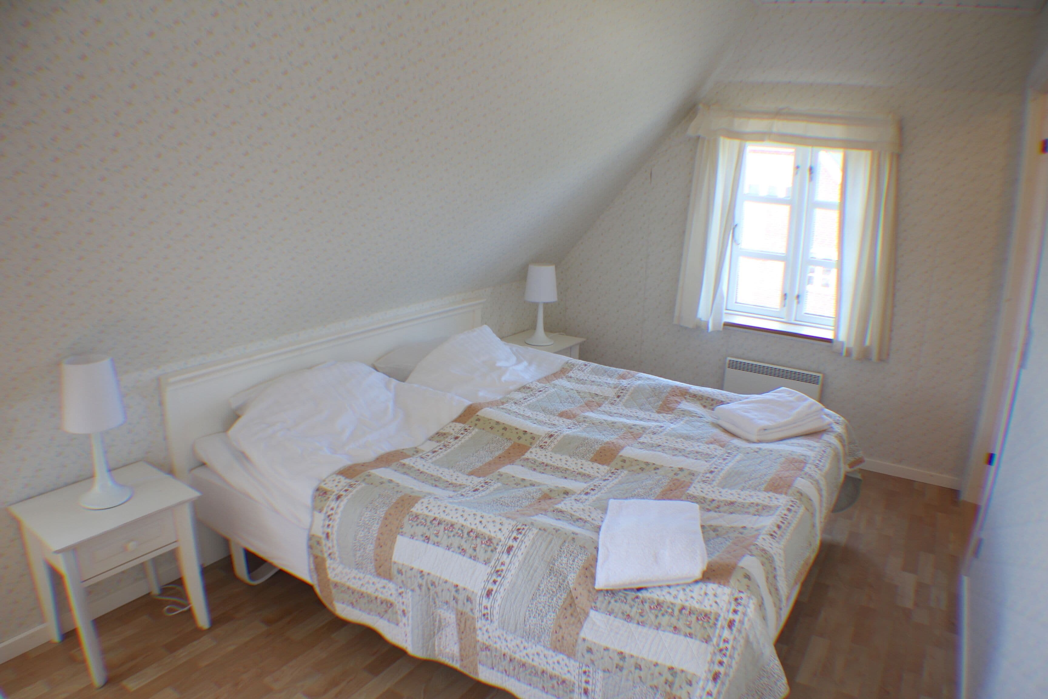 A bedroom with a bed, a nightstand, a lamp, a window, and a radiator.