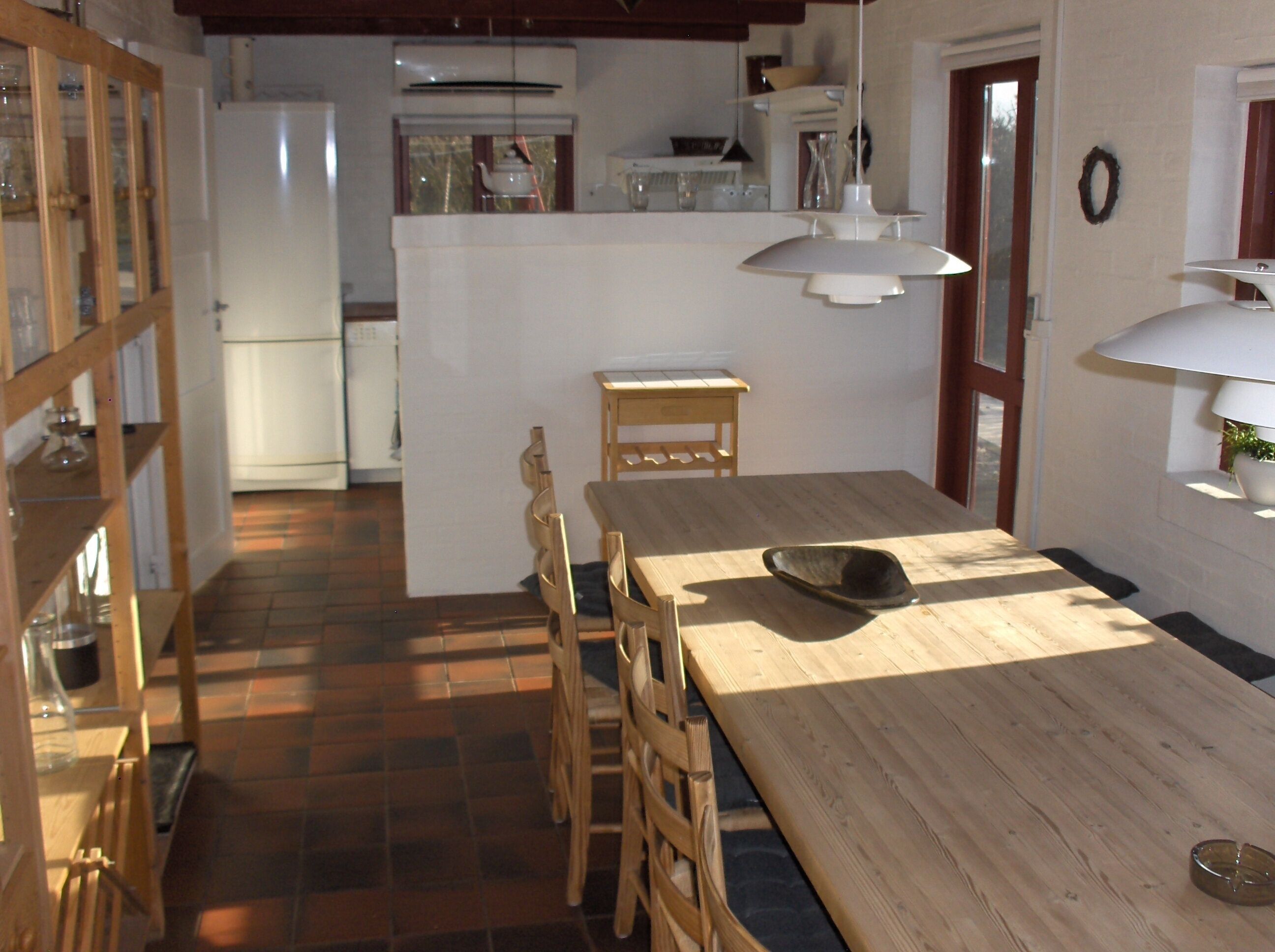 A dining area with a wooden table and chairs, a white refrigerator, and a kitchen in the background.