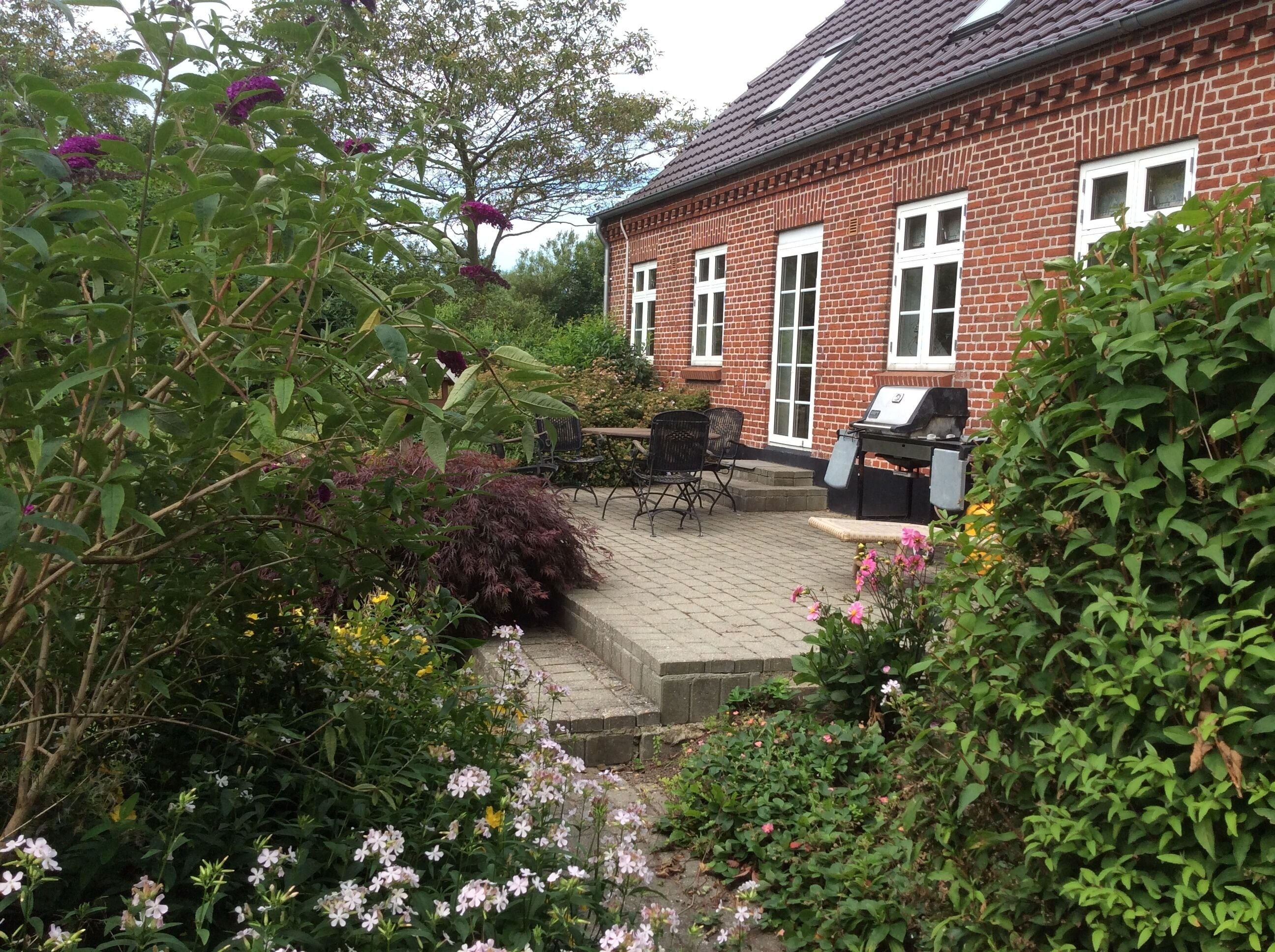 Et murstenshus med terrasse, udendørs siddepladser og grill.