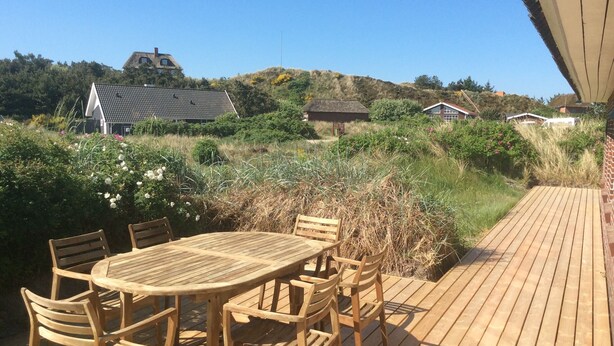 Una terraza de madera con una mesa redonda y sillas, rodeada de vegetación natural y casas al fondo.