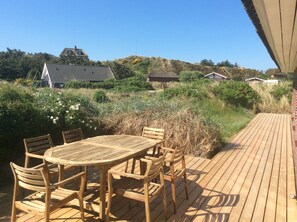 A wooden deck with a round table and chairs, surrounded by natural vegetation and houses in the background. - Klitgarden (Henne)