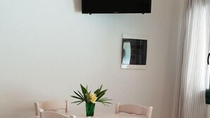 Un coin repas avec une table et des chaises blanches, une petite plante en pot et un téléviseur à écran plat fixé au mur.