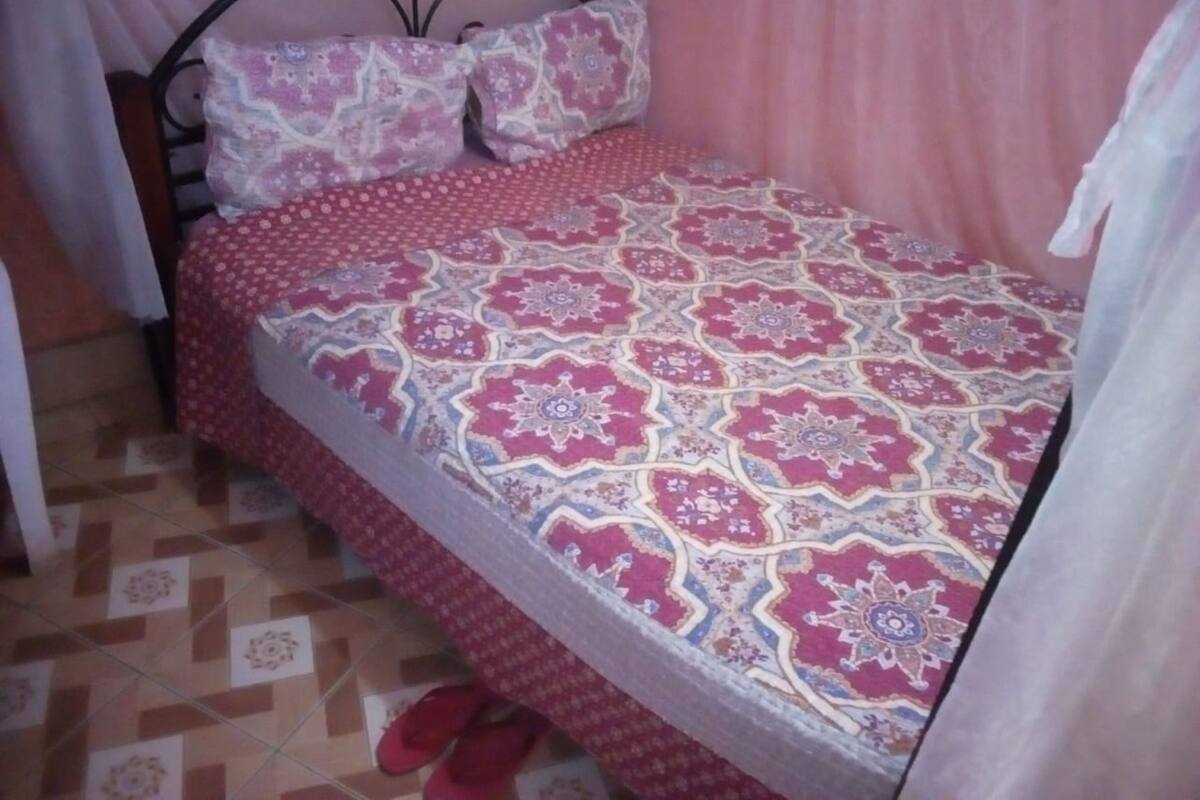 A bedroom with a patterned bedspread, a metal bed frame, and a pair of red slippers on the floor.
