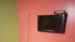 A small room with a white table, a black basket, and a television mounted on an orange wall. - Club Woodland (Matuu)