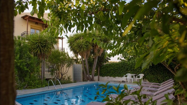 A swimming pool with lounge chairs and a small table, surrounded by greenery and a building in the background. - Villa Sofia (Faistos)