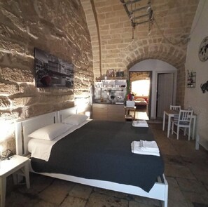 Quarto com uma cama grande, uma escrivaninha e uma cadeira. É visível uma área de cozinha através de uma porta aberta.