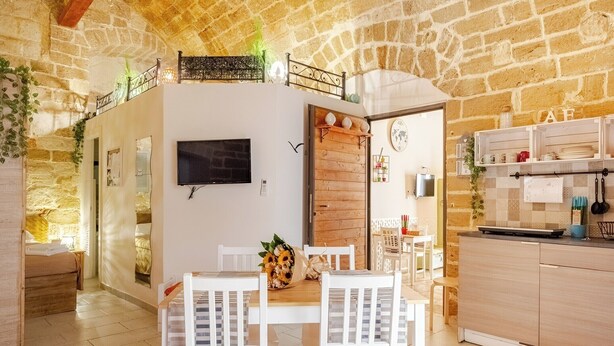 A cozy room with a stone wall, a wooden ceiling, a dining area with white chairs, a wooden table, and a kitchen with cabinets and a counter. - "LULLABY ROOM" APARTMENT BARI. (Bari)