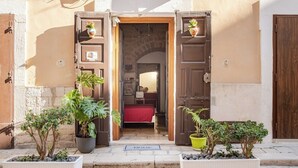 A narrow street with a building entrance featuring two open wooden doors, a red bed visible inside, and potted plants on the pavement. - "LULLABY ROOM" APARTMENT BARI. (Bari)