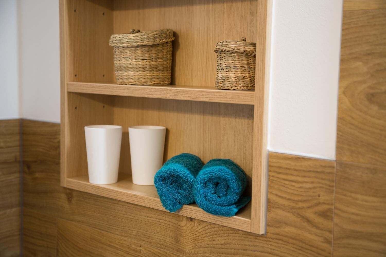 A wooden shelf with two woven baskets, three white cups, and two rolled blue towels.