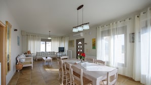 A spacious living area with a dining table, chairs, and a sofa. There is a television and a small table with decorative items. - Villa Xenophon (Faistos)
