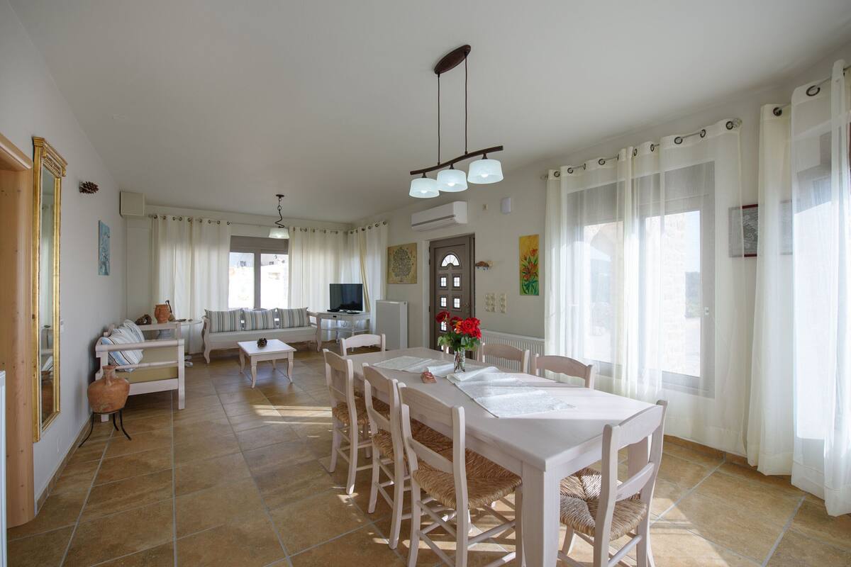 A spacious living area with a dining table, chairs, and a sofa. There is a television and a small table with decorative items.