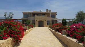 A two-story house with a stone facade, a central entrance, and a paved pathway leading to the door. - Villa Xenophon (Faistos)