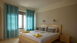 A neatly made bed with white linens and yellow towels, a wooden headboard, a bedside table with a lamp, and a window with curtains. - Villa Xenophon (Faistos)