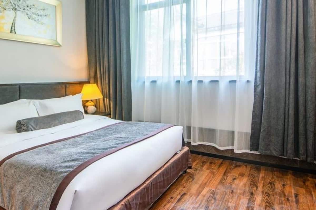 A hotel room with a large bed, a nightstand with a lamp, a painting on the wall, and a window with curtains.