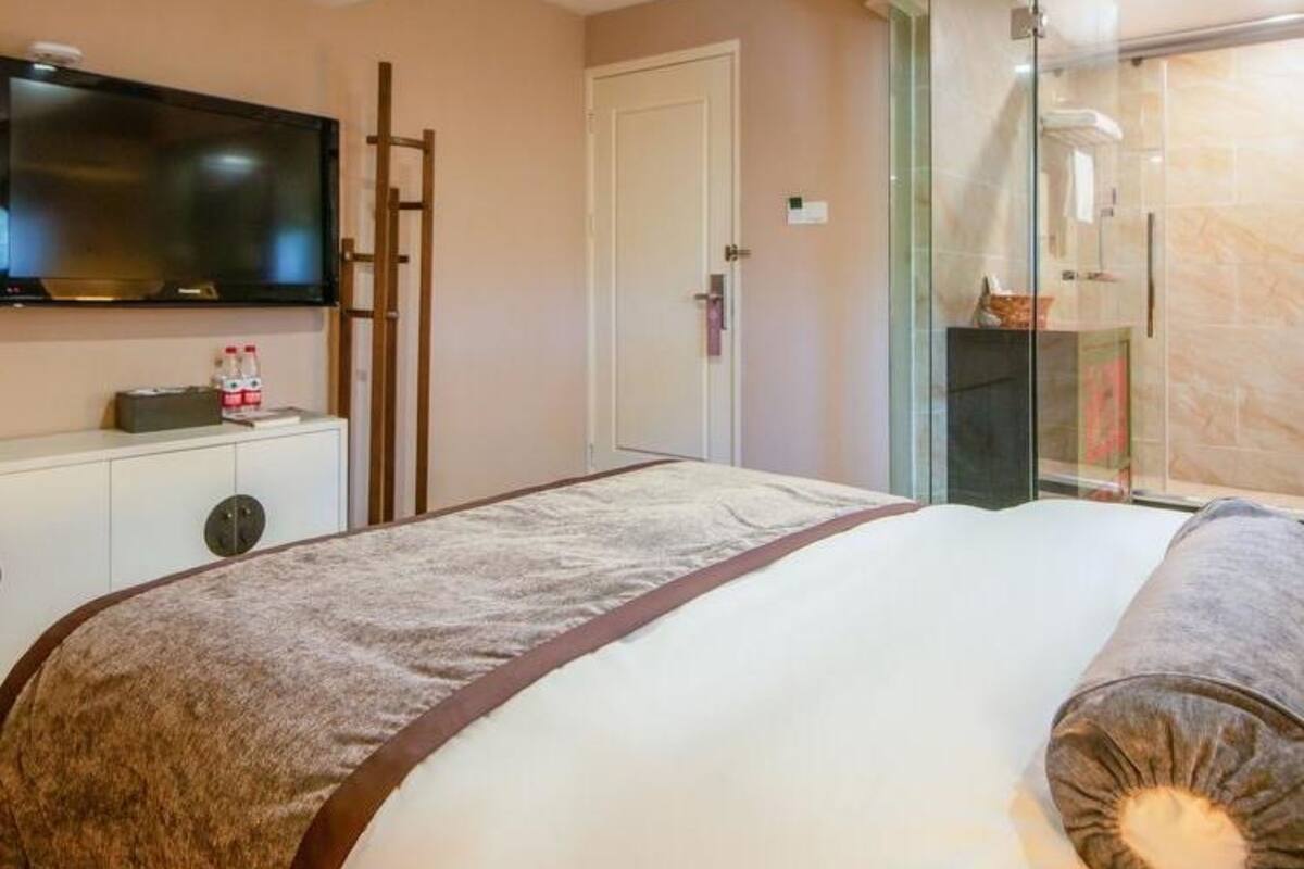 A hotel room with a bed, a TV mounted on the wall, a wardrobe, and a glass shower enclosure.