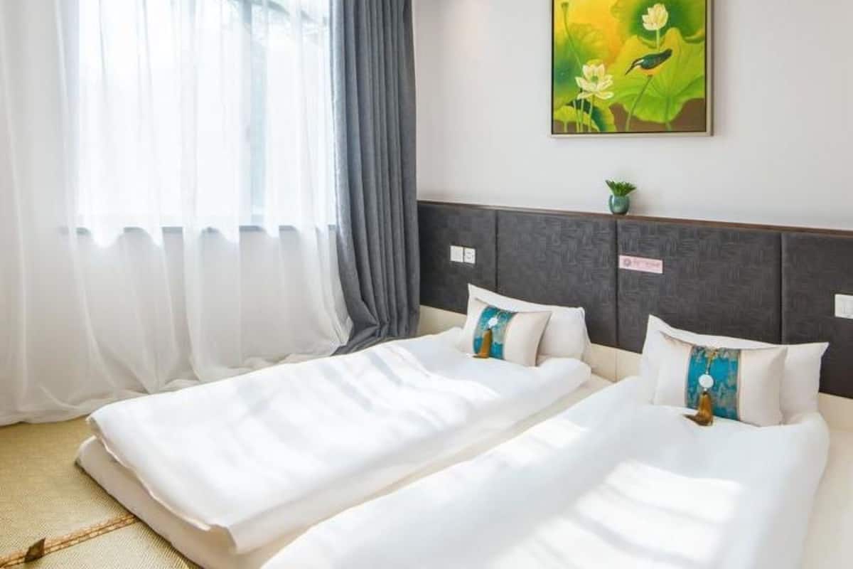 A hotel room with two beds, a large window with sheer curtains, a painting of flowers on the wall, and a small plant on the nightstand.