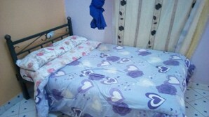 A bed with floral bedding, a pair of green slippers on the floor, and a blue garment hanging on the wall. - Dream Light Hotel (Matuu)