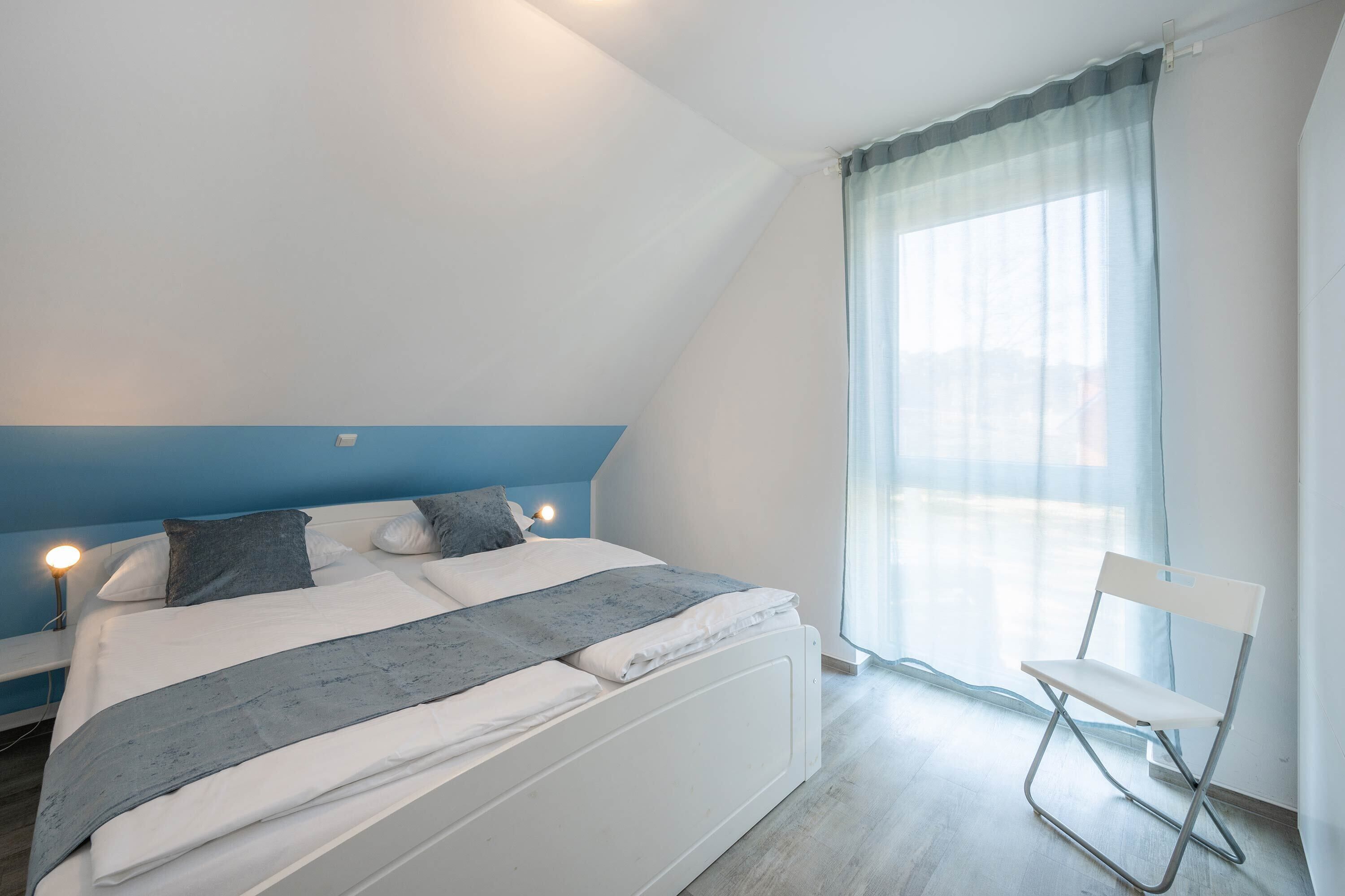 Une chambre à coucher avec un lit, une chaise, une fenêtre avec des rideaux et une table de chevet avec une lampe.