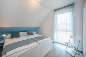 A bedroom with a bed, a chair, a window with curtains, and a nightstand with a lamp.