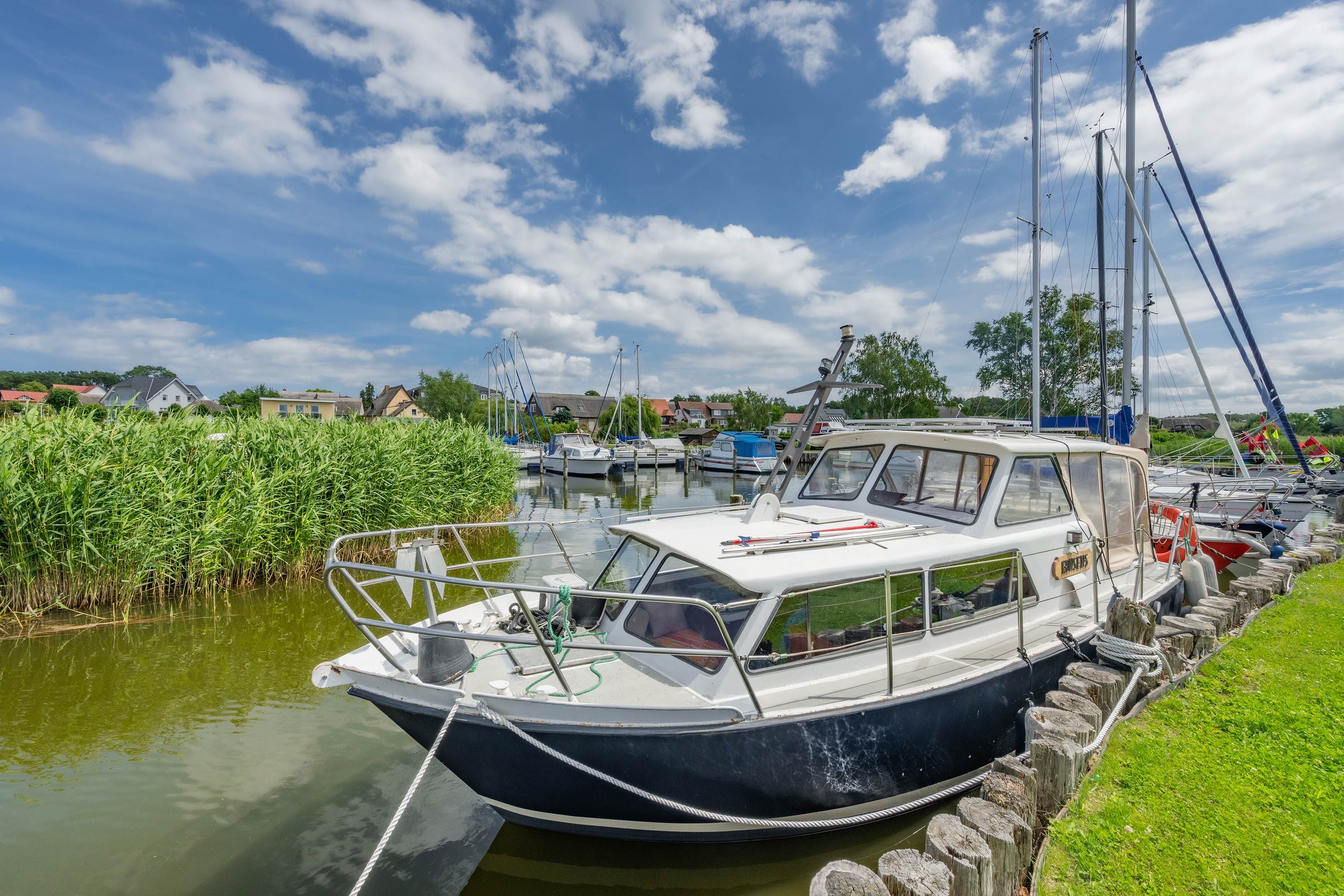 Ein Boot liegt in einer Marina an, im Hintergrund sind Häuser und ein grasbewachsener Uferstreifen zu sehen.