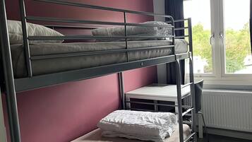 Quarto com beliche, escrivaninha, cadeira, radiador e janela com cortinas.
