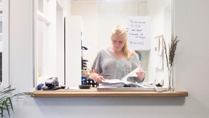 Eine Person arbeitet an einem Schreibtisch mit Computer, Unterlagen und einer Pflanze.