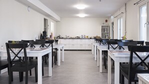 Ein moderner Essbereich mit weißen Tischen und schwarzen Stühlen, im Hintergrund eine Küche und eine Uhr an der Wand.
