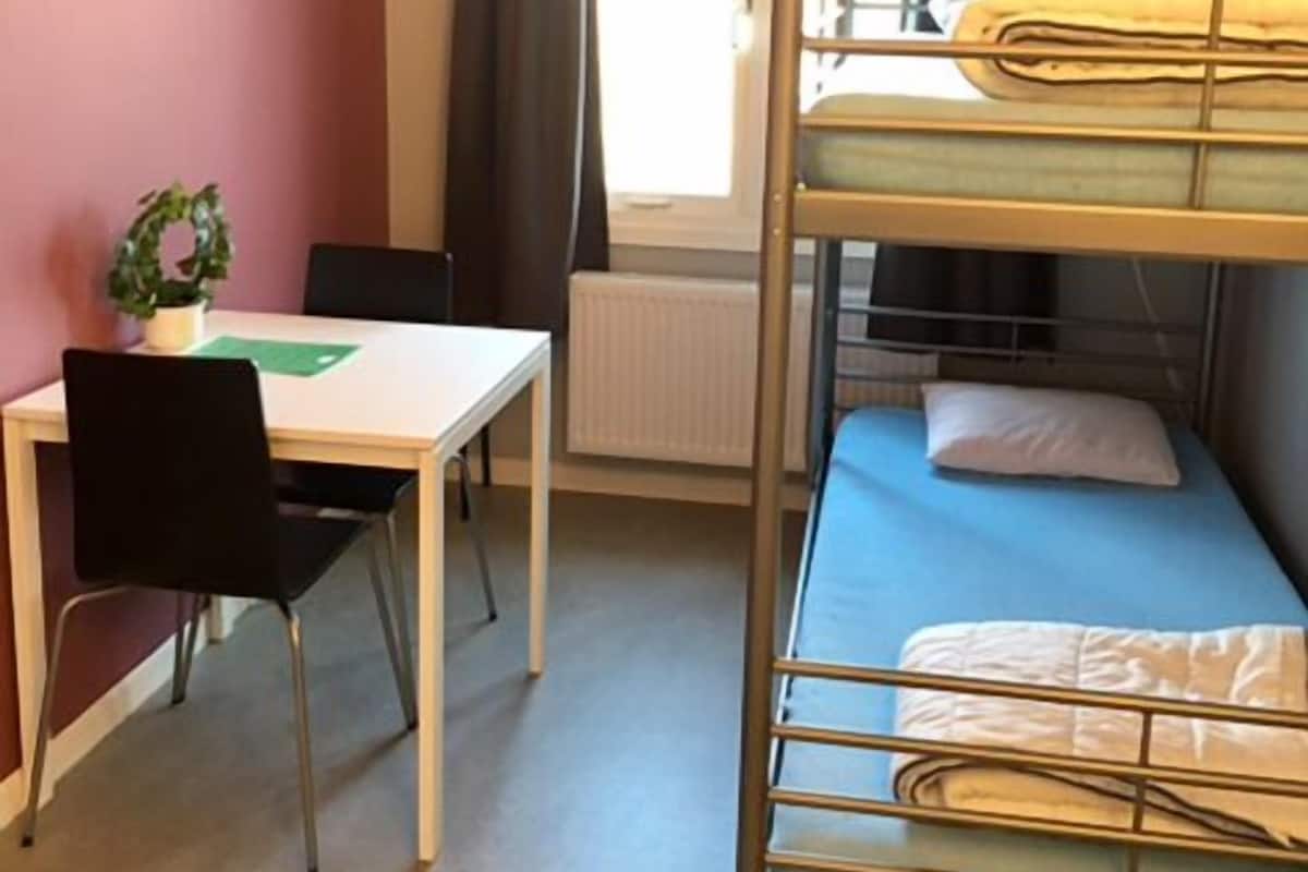 Une chambre de dortoir avec des lits superposés, un bureau, une chaise, un radiateur et une fenêtre avec des rideaux.