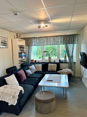 Un salon comprenant un canapé d’angle, une table basse, un petit pouf, une bibliothèque, un radiateur et un téléviseur.