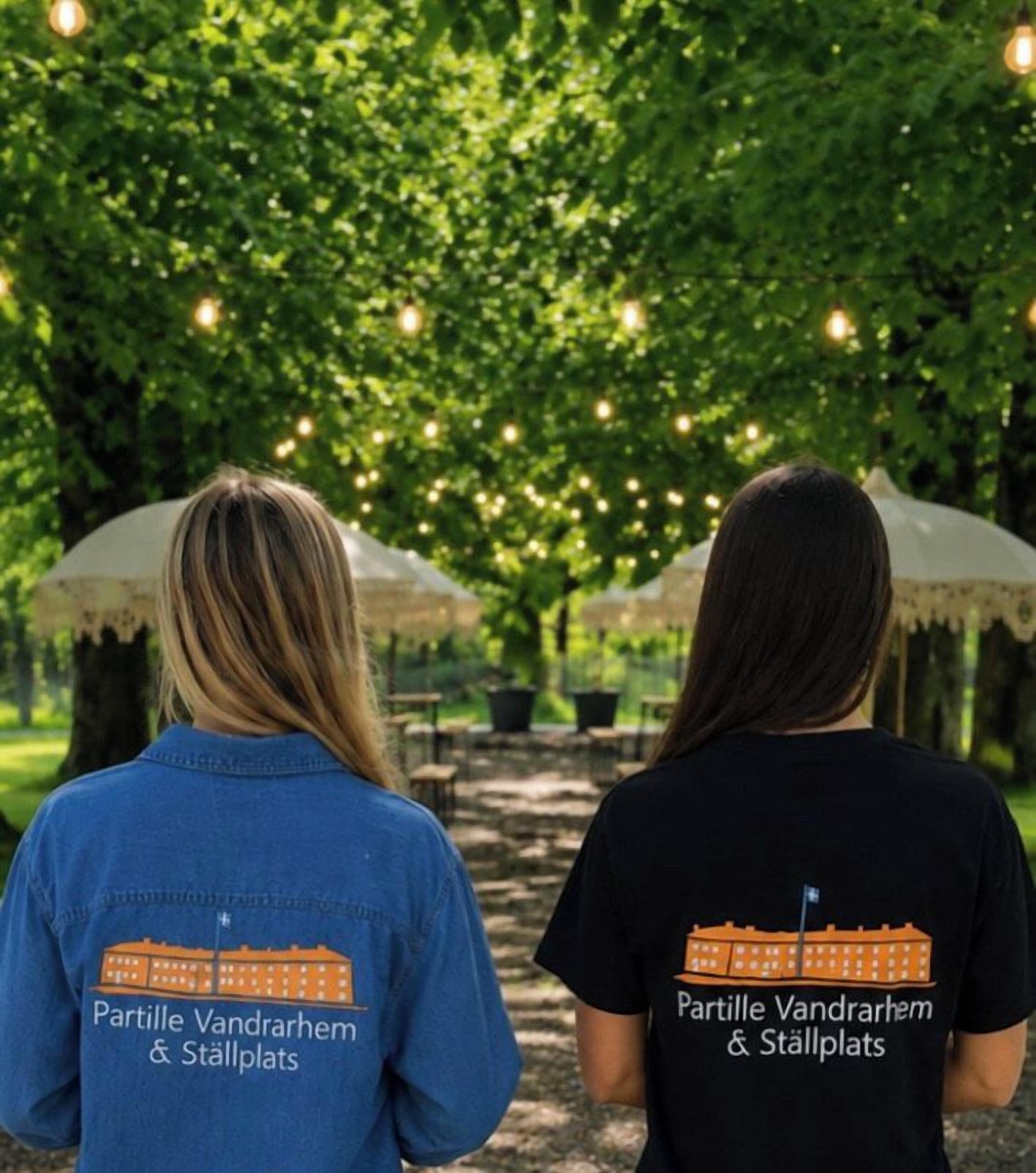 Two individuals wearing t-shirts with a building graphic and text, standing under a tree with hanging lights.