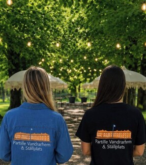 Twee personen met t-shirts met een afbeelding van een gebouw en tekst, staan onder een boom met hangende lichtjes.