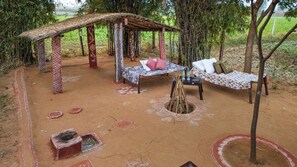 Un espace extérieur rustique aménagé avec une structure à toit de chaume, deux bancs dotés de coussins et un petit foyer.